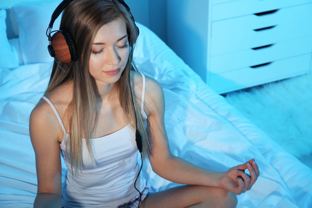 young woman meditating and listening to music while sitting on bed at home