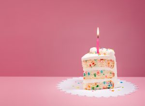 Slice of Colorful Birthday Confetti Cake with a lit candle over a pink background.