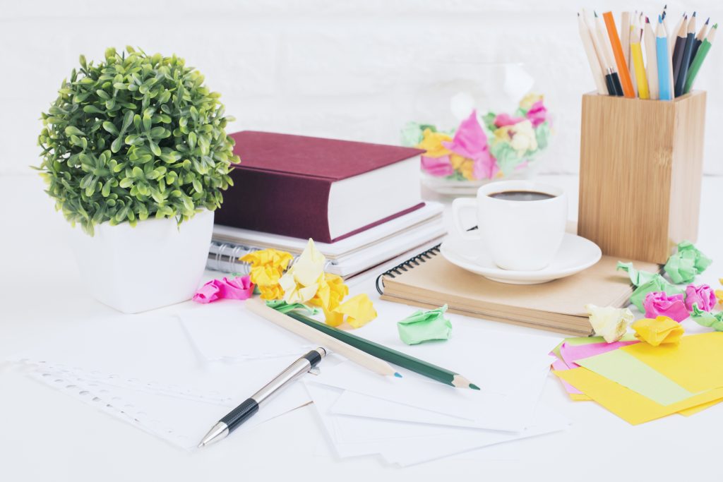 Closeup of messy office desktop with colorful crumpled paper balls, coffee cup, sationery items, decorative plant, book and other items on white brick wall background